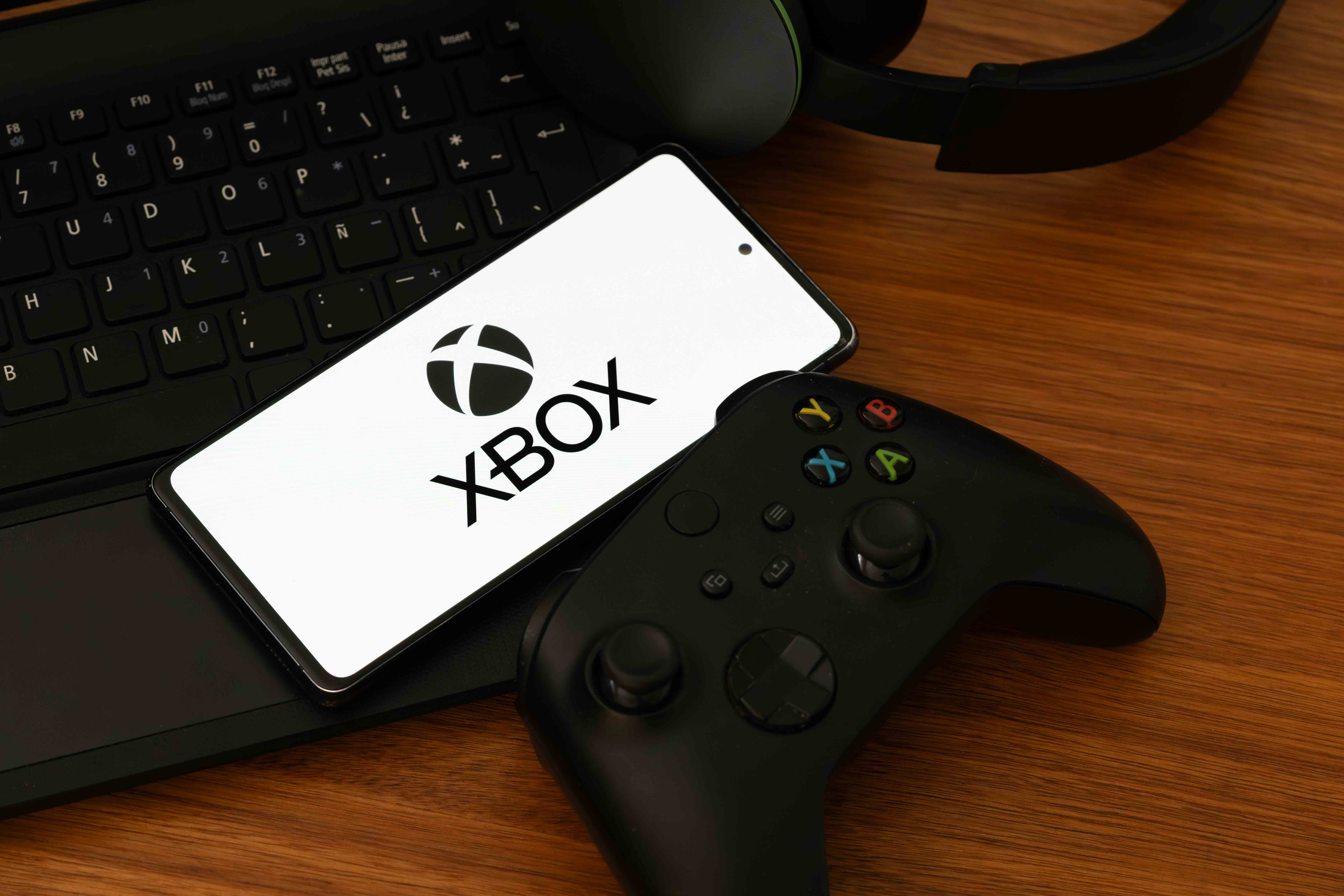 Smartphone displaying Xbox logo on screen next to game controller and laptop keyboard on wooden desk