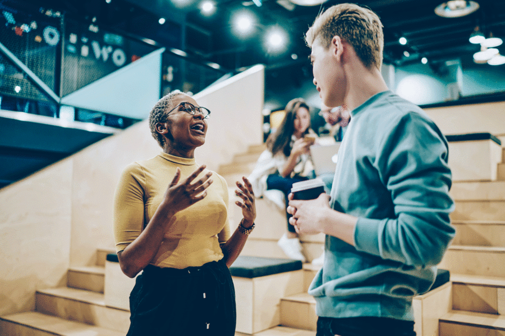 A Black woman with glasses in a yellow turtle neck speaking to an Asian man holding a coffee cup using her hands in an emotionally intelligent way