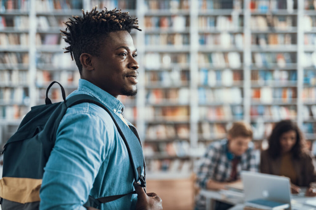 Black man in a light blue collared shirt wearing a backpack in a library.