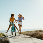 Two young women have fun with a bicycle at the beach. Woman running with friend riding a bike on boardwalk.