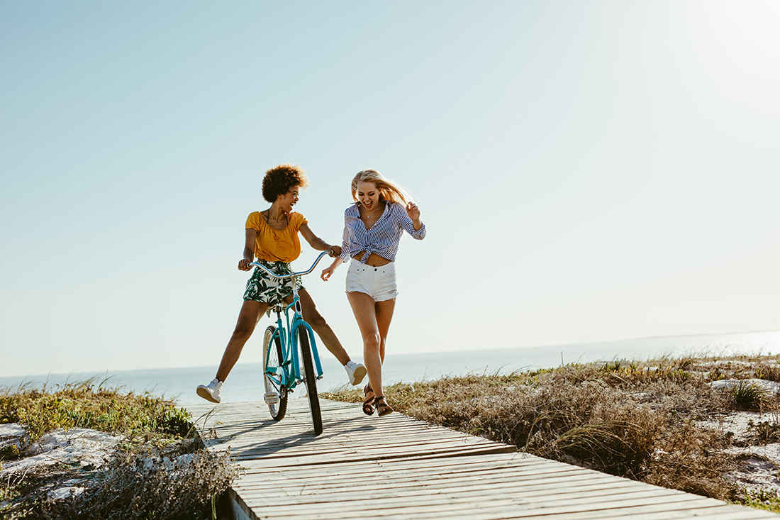 Two young women have fun with a bicycle at the beach. Woman running with friend riding a bike on boardwalk.