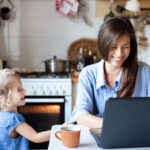 Working mom works from home office. Happy mother and daughter smiling. Successful woman and cute child using laptop. Freelancer workplace in cozy kitchen. Female business. Lifestyle authentic moment.