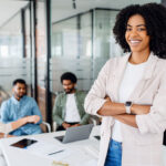 woman with a cheerful smile stands with arms crossed on the forefront in a modern office setting with her colleagues collaborating in the background,