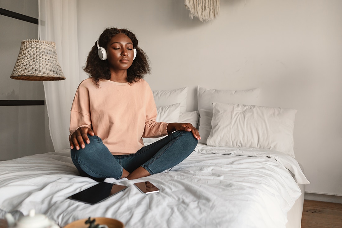 Online Meditation. Woman Meditating With Eyes Closed Wearing Headphones Sitting In Lotus Position, Listening To Yoga Instructor Via Digital Tablet Relaxing In Bedroom At Home
