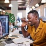 A Black man waves to his colleague on a video call from his office