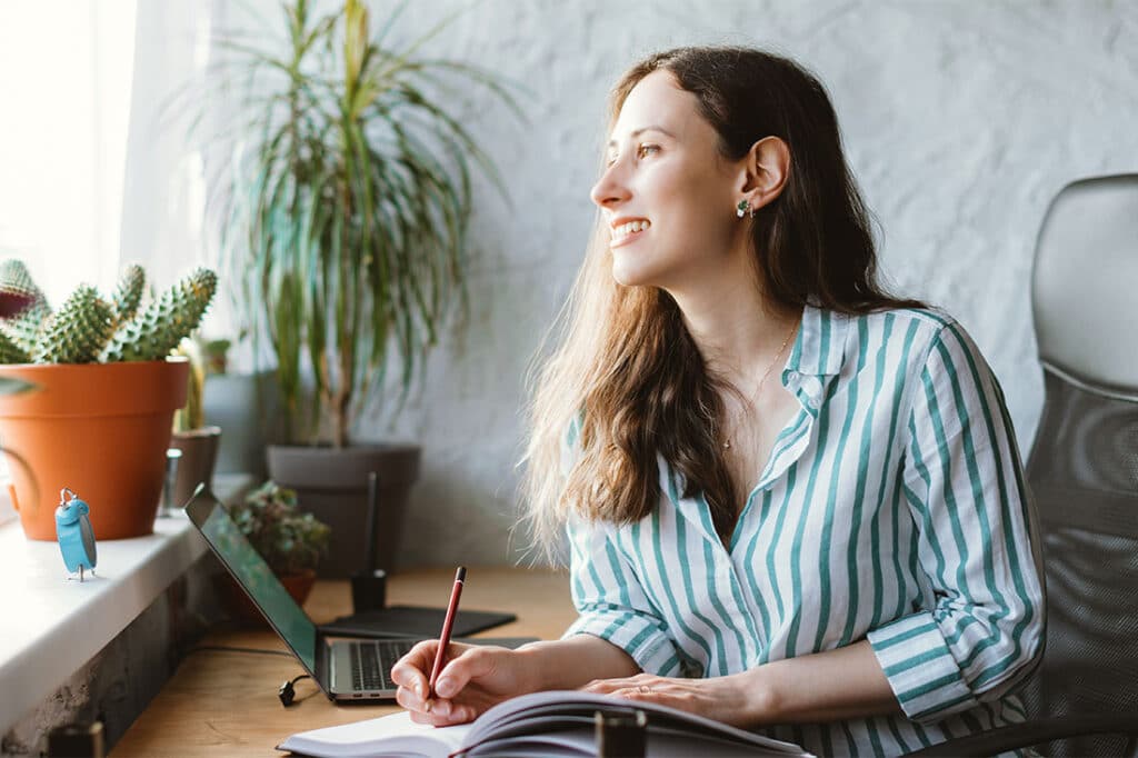 woman is looking through window while writing in journal at her work desk