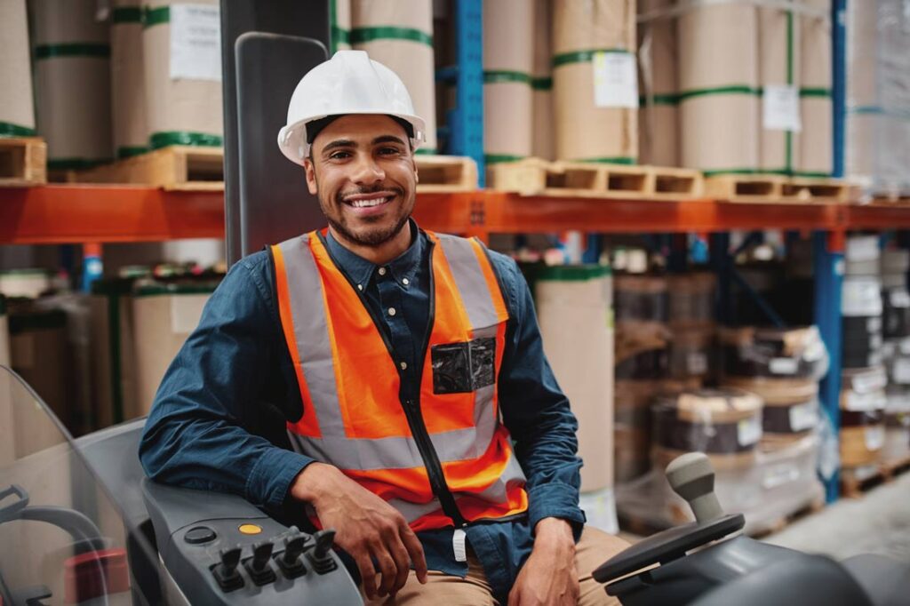 a young stockroom employee participating in the Big Stay smiles while wearing a hard hat and orange vest