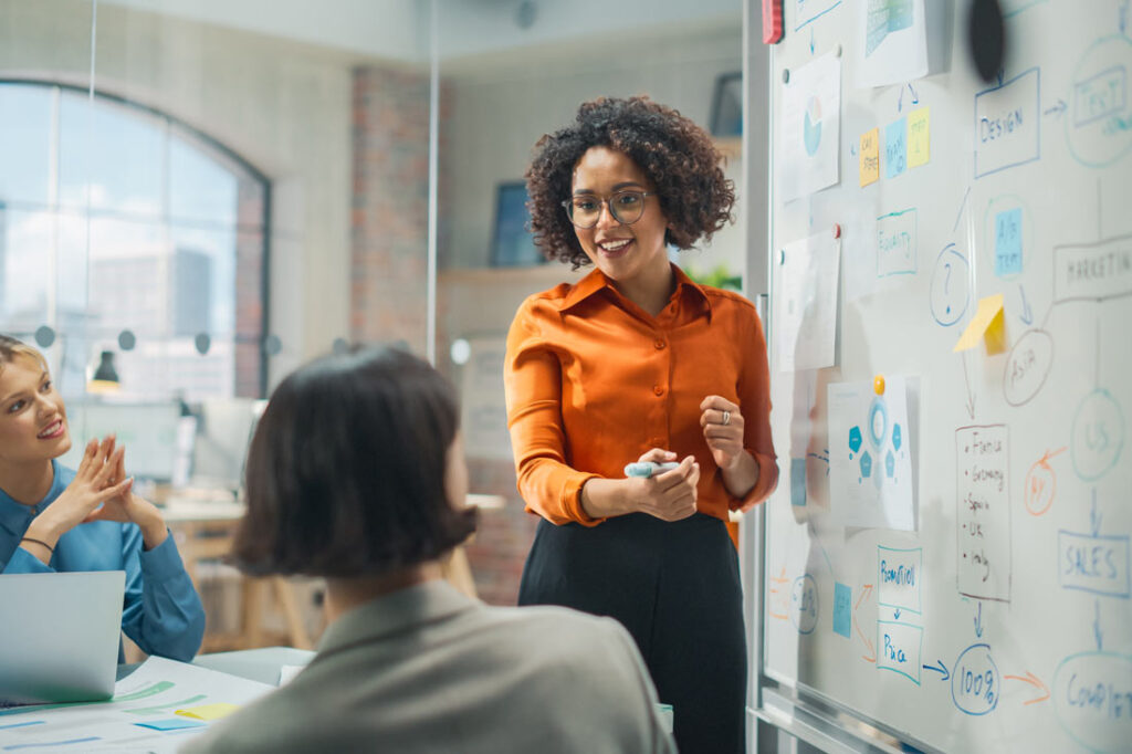 woman standing in front of a white board talks with coworkers in a conference room about inclusive marketing