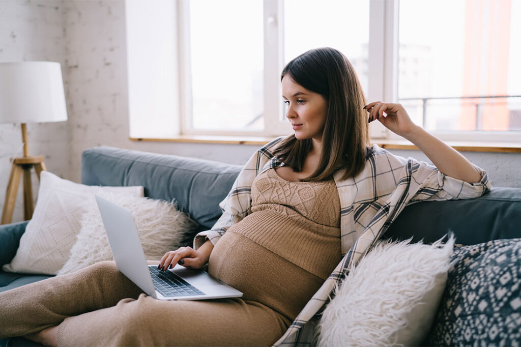 young pregnant female freelancer browsing and looking at screen of laptop while relaxing on sofa