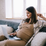 young pregnant female freelancer browsing and looking at screen of laptop while relaxing on sofa