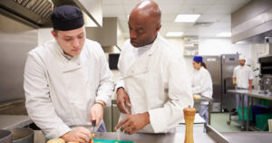 a middle-aged chef watches as an intern cuts vegetables in a restaurant kitchen, an example of working with interns