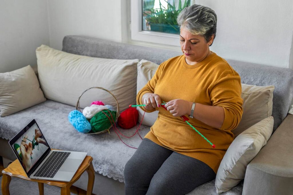 woman crocheting on a couch while watching a tutorial on her laptop
