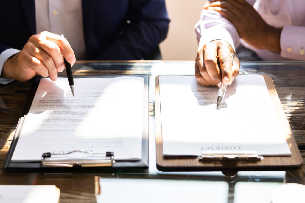 two hands holding pens, each hovering over paperwork to decide between an agreement vs contract