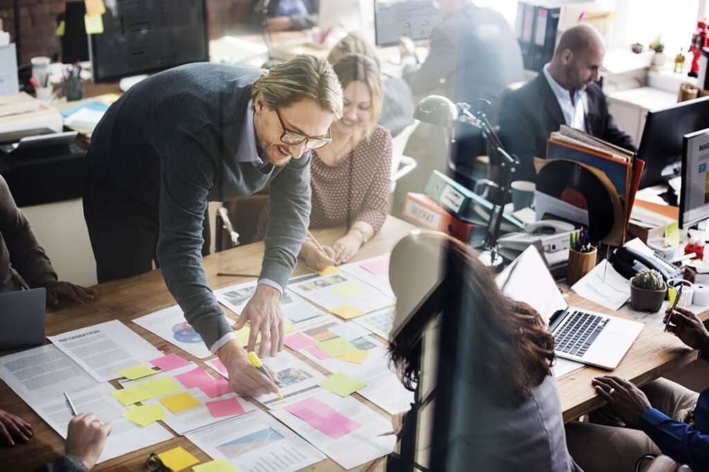 Photograph of a white man with thick rimmed glasses and two women working over a desk with lots of papers and post it notes