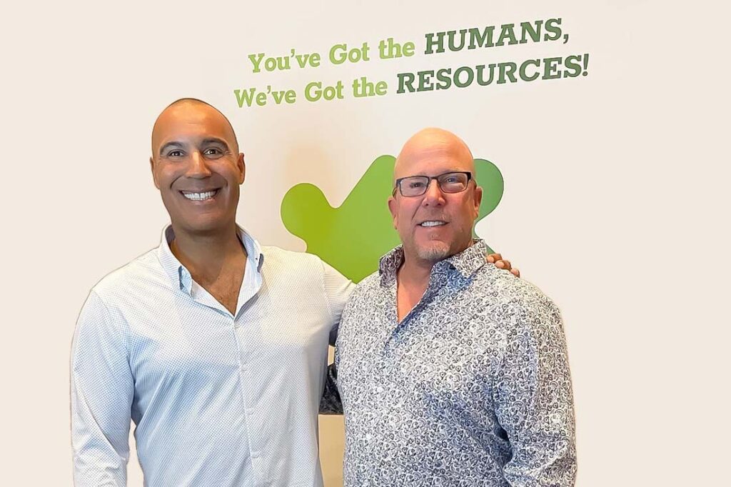 Photograph of the founders of Puzzle HR, a tall Black bald man smiling to the left and a shorter white man with glasses also smiling to the right. On the wall behind them text reads: "You've got the humans, we've got the resources."