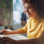 Woman of color with short curly hair in a yellow turtleneck smiles while looking at her computer and touching her keyboard and mouse.