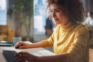 Woman of color with short curly hair in a yellow turtleneck smiles while looking at her computer and touching her keyboard and mouse.