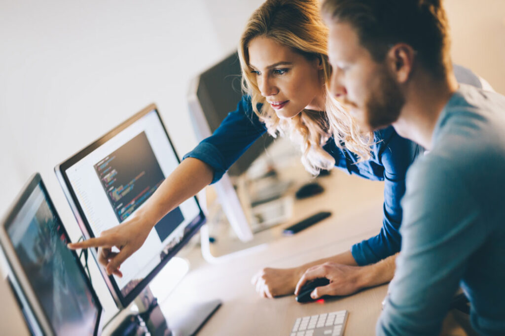 Photograph of a dark skinned woman explaining something to a light skinned man with a beard in front of a computer screen