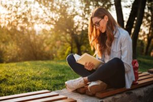 Photograph of a white woman with long straight brown hair reading books on emotional intelligence at sunset. She is sitting outside cross legged on a wooden bench