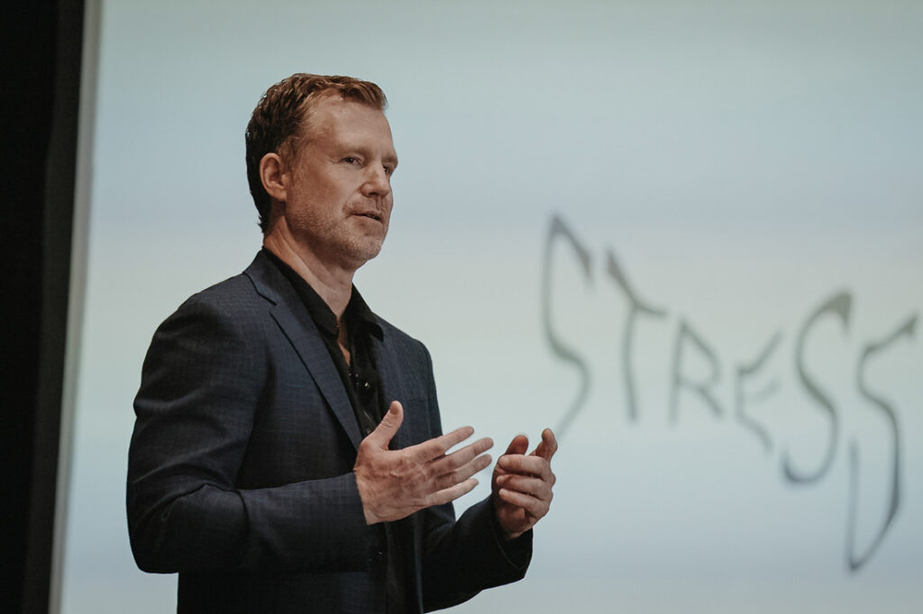Photograph of a white man in a grey suit speaking with his hands in front of a white board that says "stress"