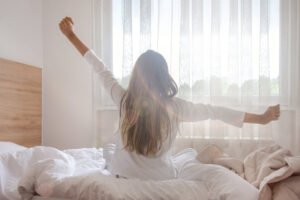 Photograph of a woman with long brown hair from behind sitting on her bed in her pajamas stretching her arms in front of a bright window to demonstrate she is a morning person