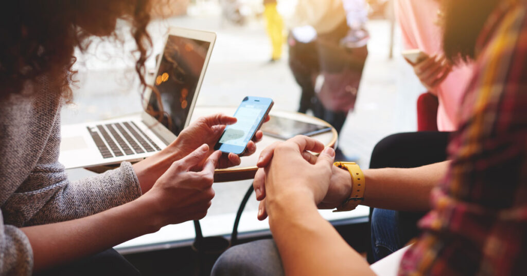 Close-up photograph of a woman holding her smart phone and showing a man in front of a laptop to represent people learning how to use AI to make money