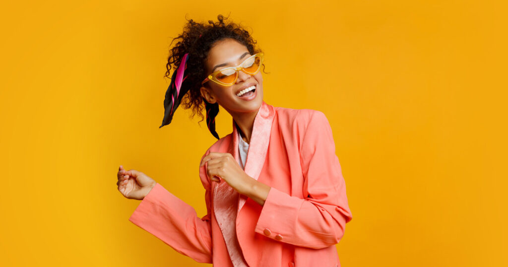 Photograph of an African American woman smiling and dancing in a salmon pink suit and yellow sunglasses to represent dopamine dressing