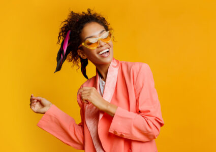 Photograph of an African American woman smiling and dancing in a salmon pink suit and yellow sunglasses to represent dopamine dressing
