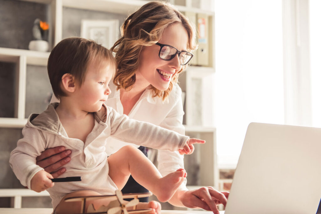 A white woman with blonde hair and glasses working from home with her toddler son pointing at the computer to show lazy girl jobs are more about flexibility than laziness
