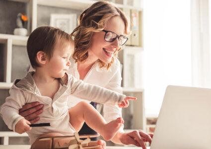 A white woman with blonde hair and glasses working from home with her toddler son pointing at the computer to show lazy girl jobs are more about flexibility than laziness