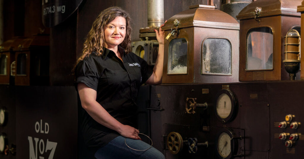 Photograph of Lexie Phillips, Jack Daniel's first female assistant distiller. She is a white woman with long curly blond hair in a black shirt, smiling in front of an old fashioned piece of distilling equipment