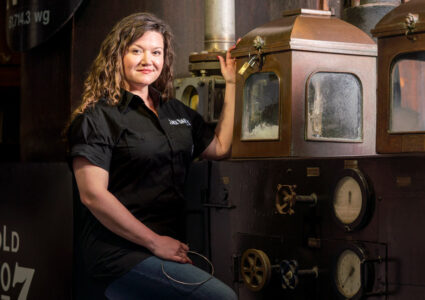Photograph of Lexie Phillips, Jack Daniel's first female assistant distiller. She is a white woman with long curly blond hair in a black shirt, smiling in front of an old fashioned piece of distilling equipment