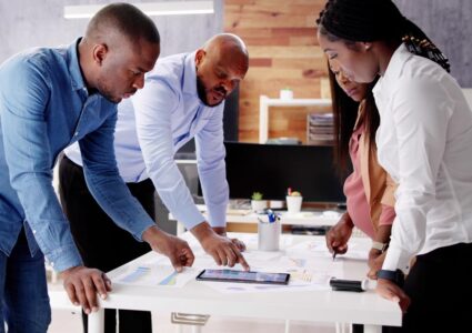 Photograph of 4 Black business people leaning over a conference table and studying a marketing swot analysis