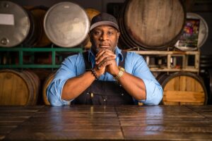 Bertony Faustin Oregons First Black Winemaker Abbey Creek Vineyard founder sitting at a wooden table in overalls in front of wine barrels