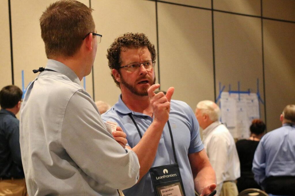 Brandon Brown owner of Continuous Coaching Commitment speaking to a man at a conference explaining why the Improvement Kata and Training Within Industry Techniques are often used together