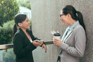 Two professionally dressed Asian women in a workplace friendship chatting outside work and smiling showing the benefits of work friends