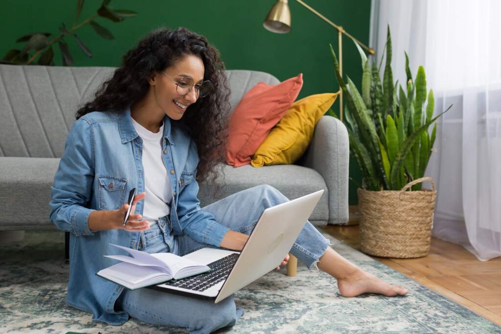 Young Latina woman sitting on the floor in her living room with computer on her leg smiling and taking a personal development course