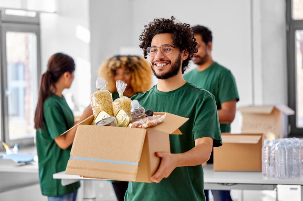 Young man holding a box of groceries and smiling because he's volunteering on Giving Tuesday 2023