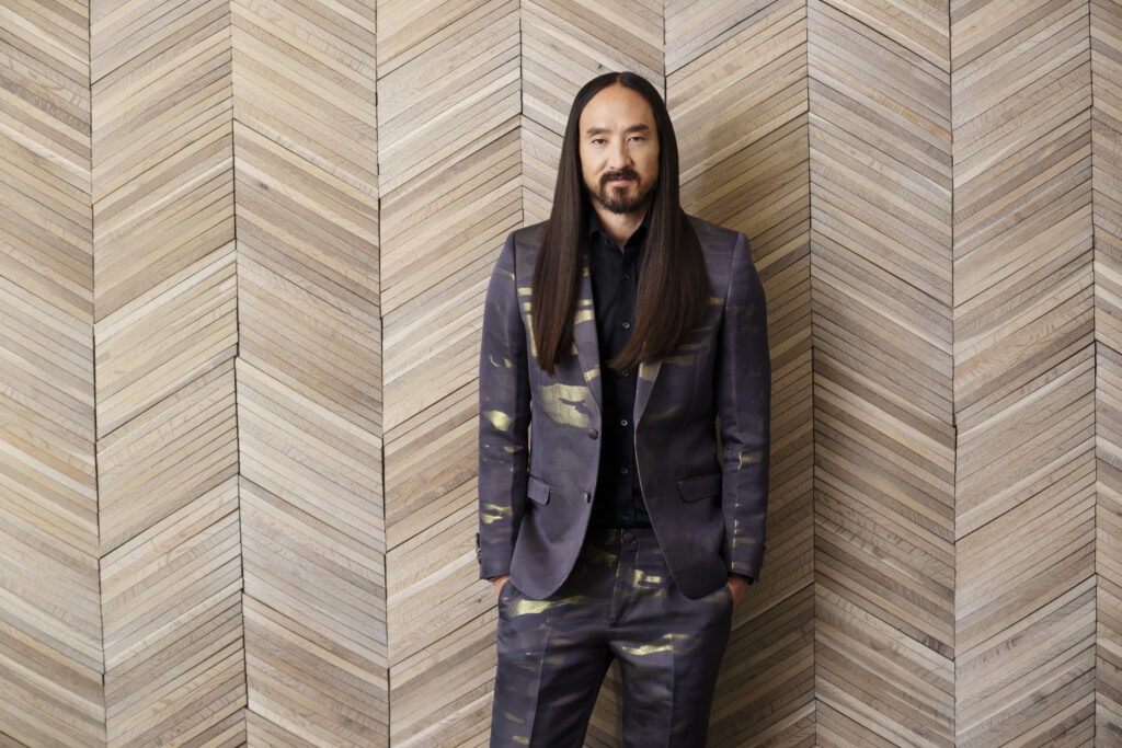 Steve Aoki in a suit posing against a herringbone-patterned wall. Photo by Eric Jamison Studio