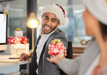 Happy male employee wearing a Santa Claus hat giving his coworker the best ten dollar gift ideas
