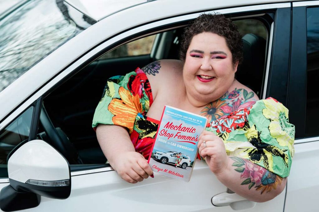 Chaya Milchtein leaning out a car window holding her new book, Mechanic Shop Femme's Guide to Car Ownership: Uncomplicating Cars for All of Us