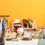 A table full of miscellaneous garage sale items including records, hats, and dishes next to a rack of clothes