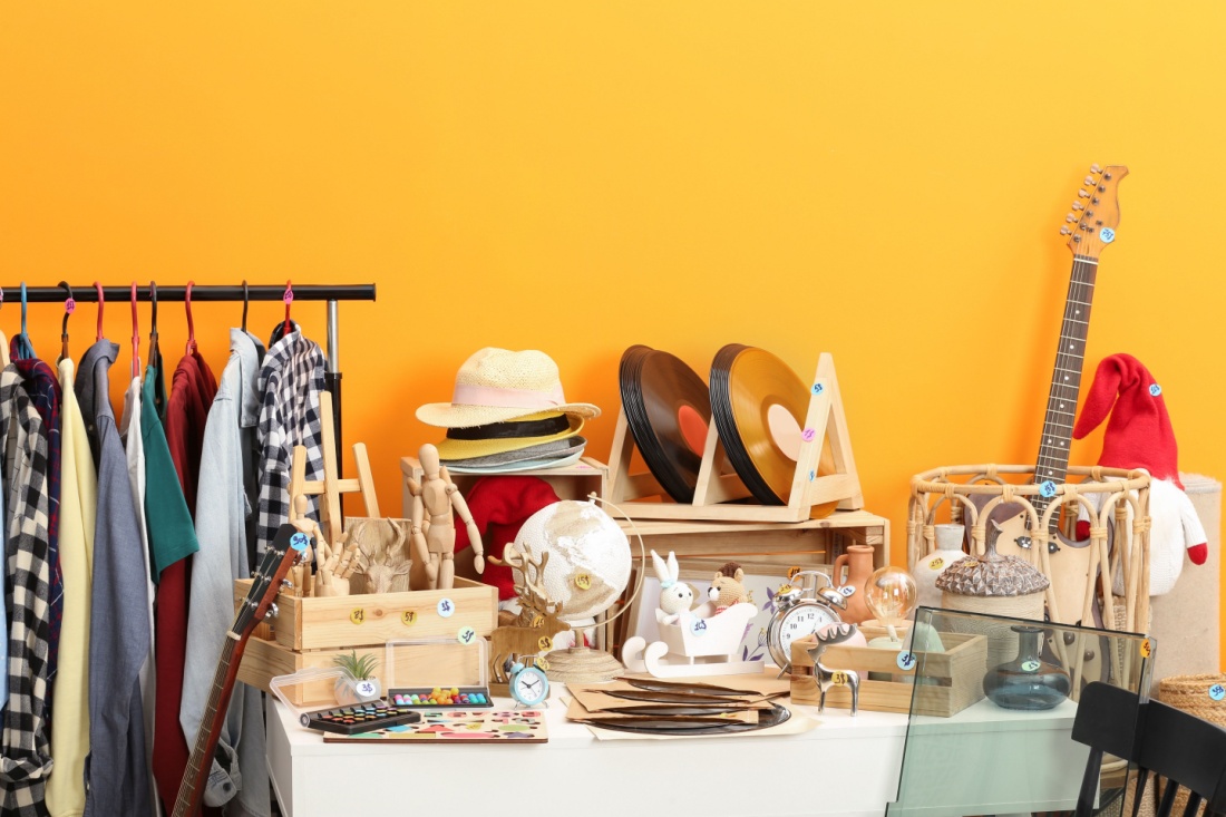 A table full of miscellaneous garage sale items including records, hats, and dishes next to a rack of clothes