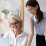 young businesswoman help middle-aged coworker with computer job at workplace