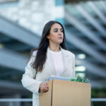 Recently laid off employee carrying cardboard box with her belongings, leaving office building after layoff, reflecting economic challenges of corporate dismissal and unemployment