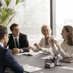 Positive elder and Gen Z coworkers laughing on corporate meeting, discussing creative ideas, brainstorming, having fun, sitting at conference table. Mature mentor, coach training employees
