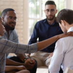 A support group, all in professional dress. A woman puts her hand on a man's shoulder to comfort him.