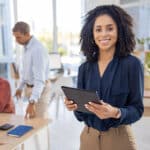 black woman holding tablet and smiling in office environment and people working in backdrop