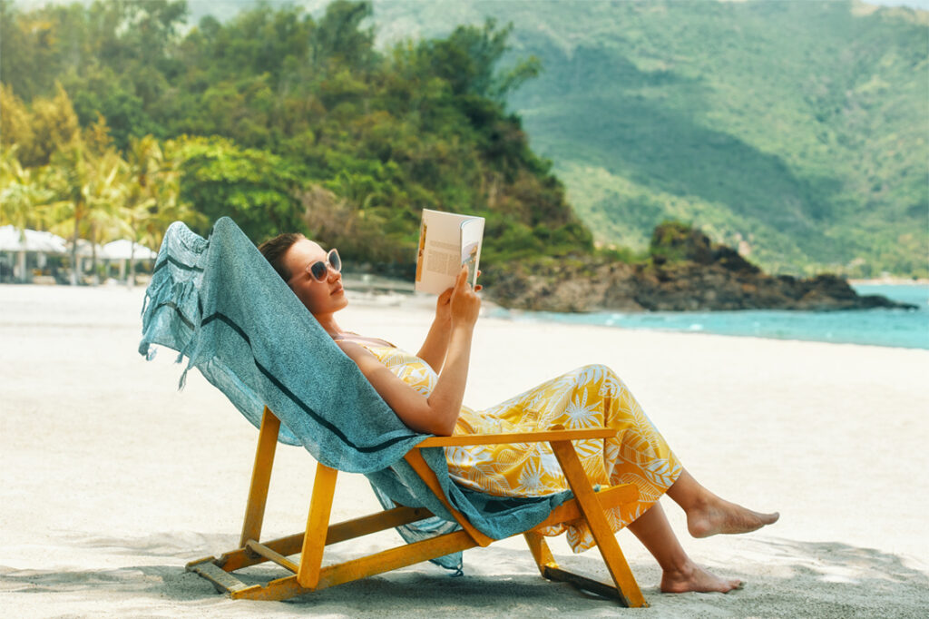 A woman sits on the beach and reads a book by the water