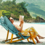 A woman sits on the beach and reads a book by the water